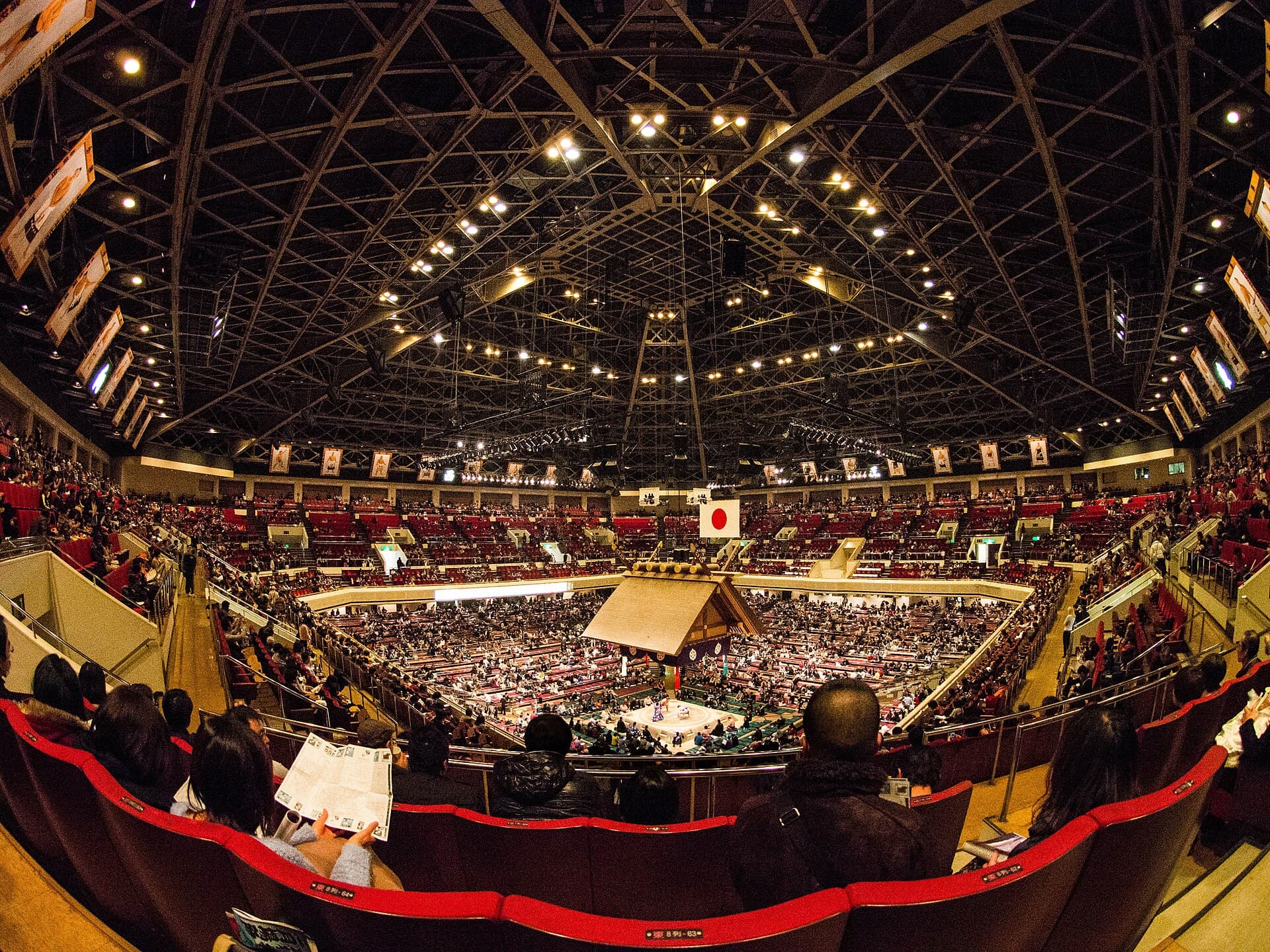 Vue plongeante de l'interieur du Ryogoku Kokugikan pendant le tournoi de janvier 2014 : le cercle d'argile et son toit de sanctuaire suspendu au centre, entoures des loges carrees sur tatami et des gradins combles de spectateurs