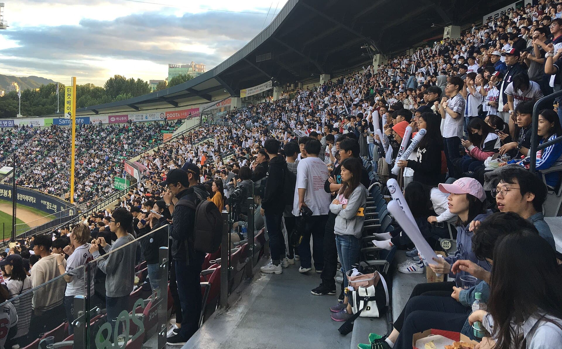 Tribunes pleines du stade de baseball de Jamsil a Seoul en soiree : des milliers de supporters debout, batons gonflables blancs a la main, chantant face au terrain pendant un match de la ligue coreenne