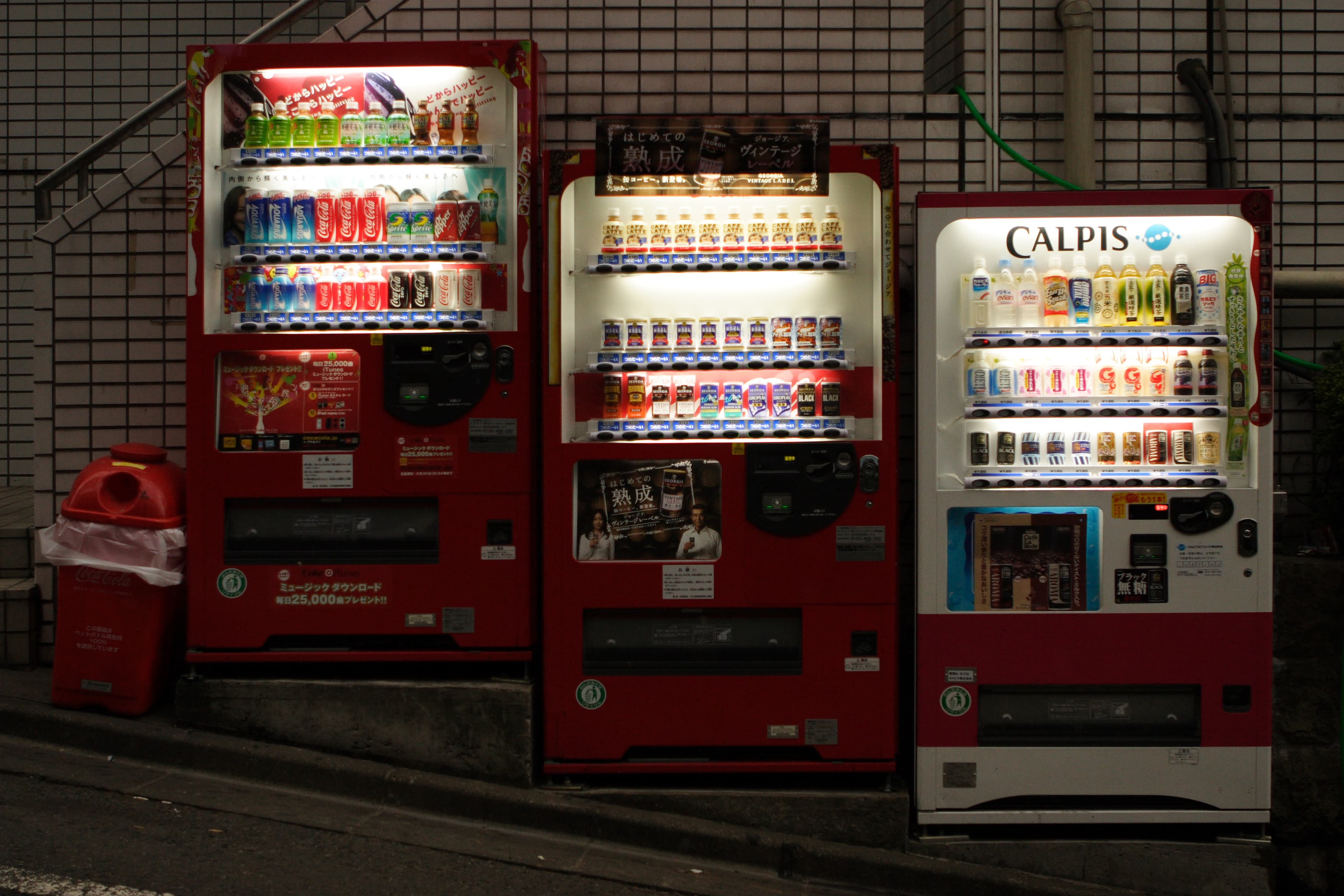 Trois distributeurs automatiques de boissons alignés le long d’une rue japonaise, éclairés la nuit.