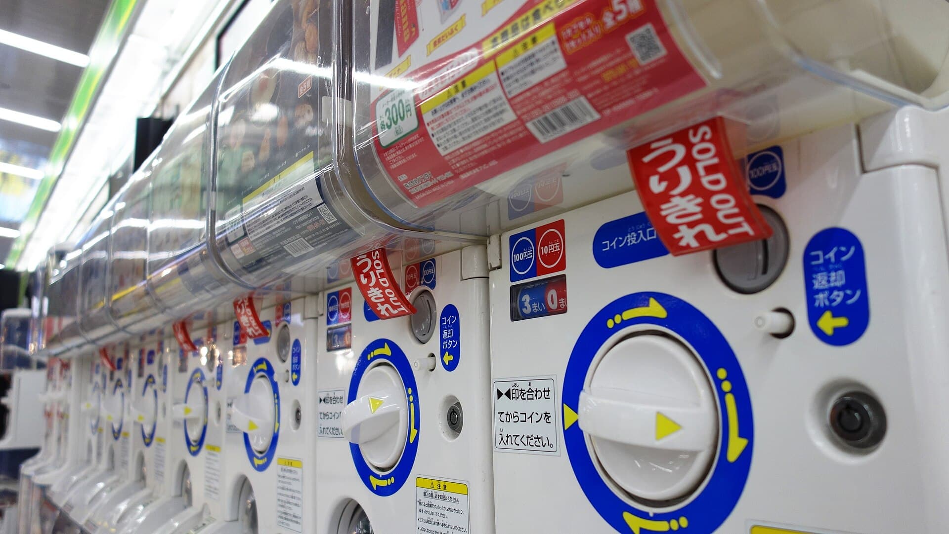 Rangee de distributeurs de capsules japonais avec leurs manivelles blanches et bleues, plusieurs portant une etiquette rouge indiquant qu'ils sont epuises
