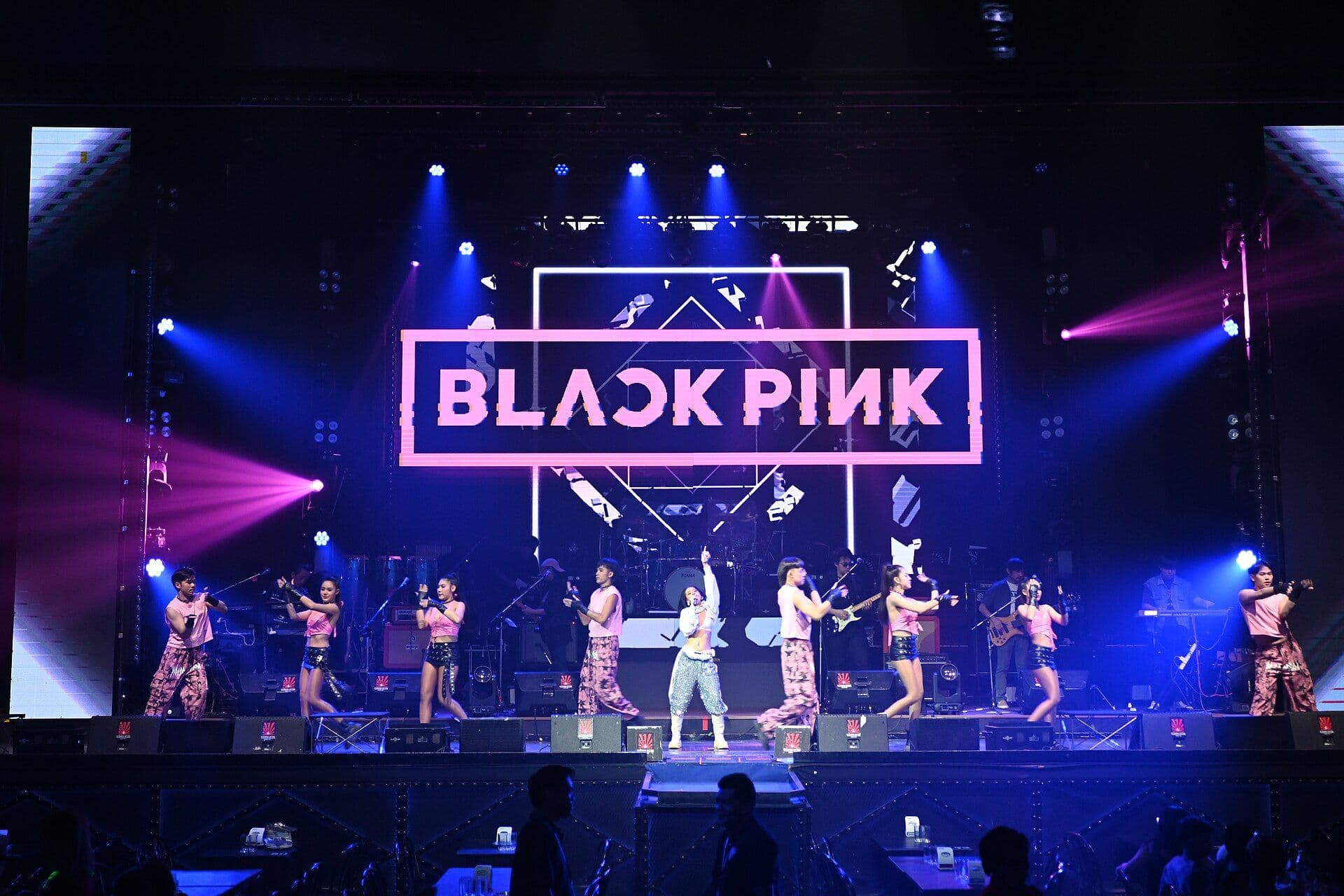 Scène de concert K-pop éclairée de rose et de bleu, danseurs devant une enseigne lumineuse.
