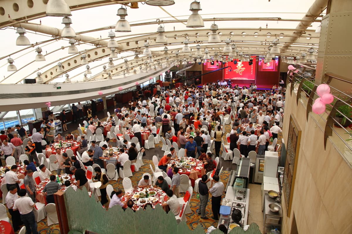Grande salle de banquet chinoise vue depuis une mezzanine : des dizaines de tables rondes garnies de plats, les convives debout entre les chaises et une scene rouge portant le caractere du bonheur double au fond de la salle