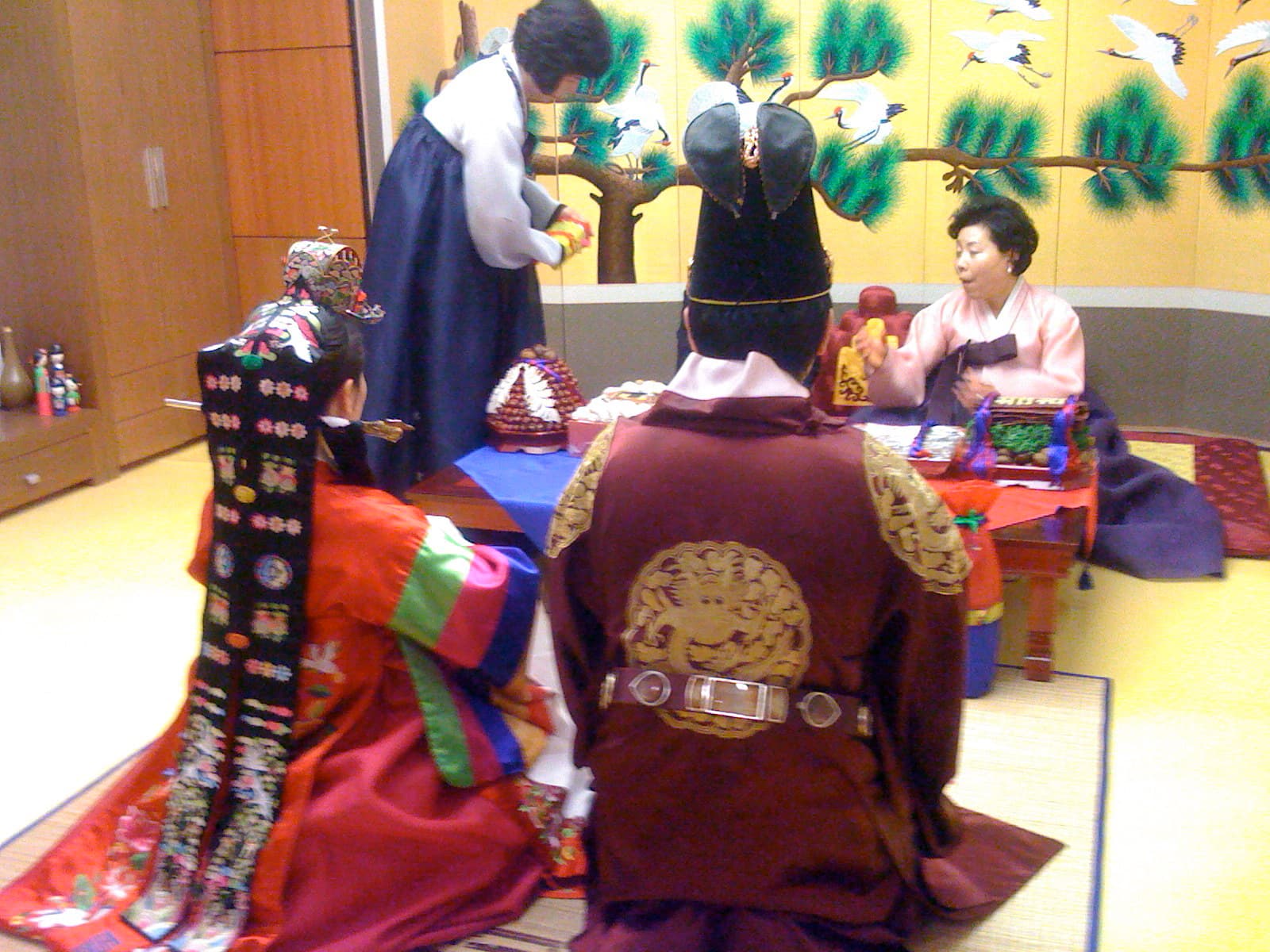 Ceremonie du pyebaek dans une salle de mariage coreenne : les maries en hanbok de ceremonie, la mariee coiffee du jokduri et le marie en costume d'officier de cour, agenouilles devant une table basse ou la belle-mere lance des jujubes, un paravent brode de pins et de grues au fond