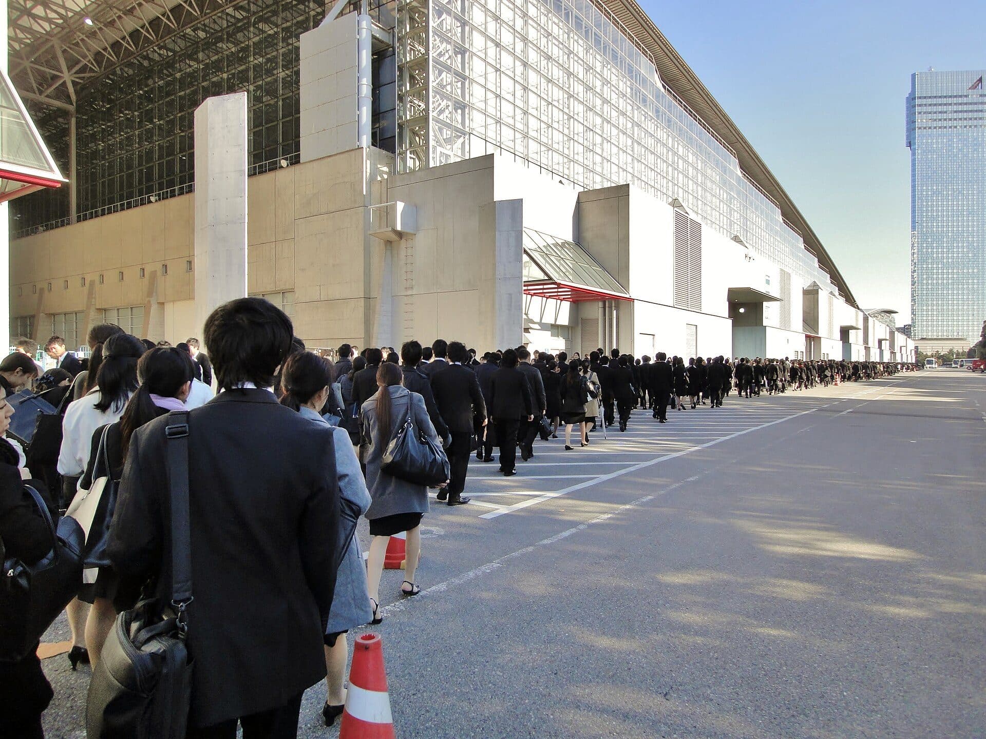 File d'étudiants japonais en costume de recrutement noir devant un centre de congrès, pendant un forum d'entreprises.