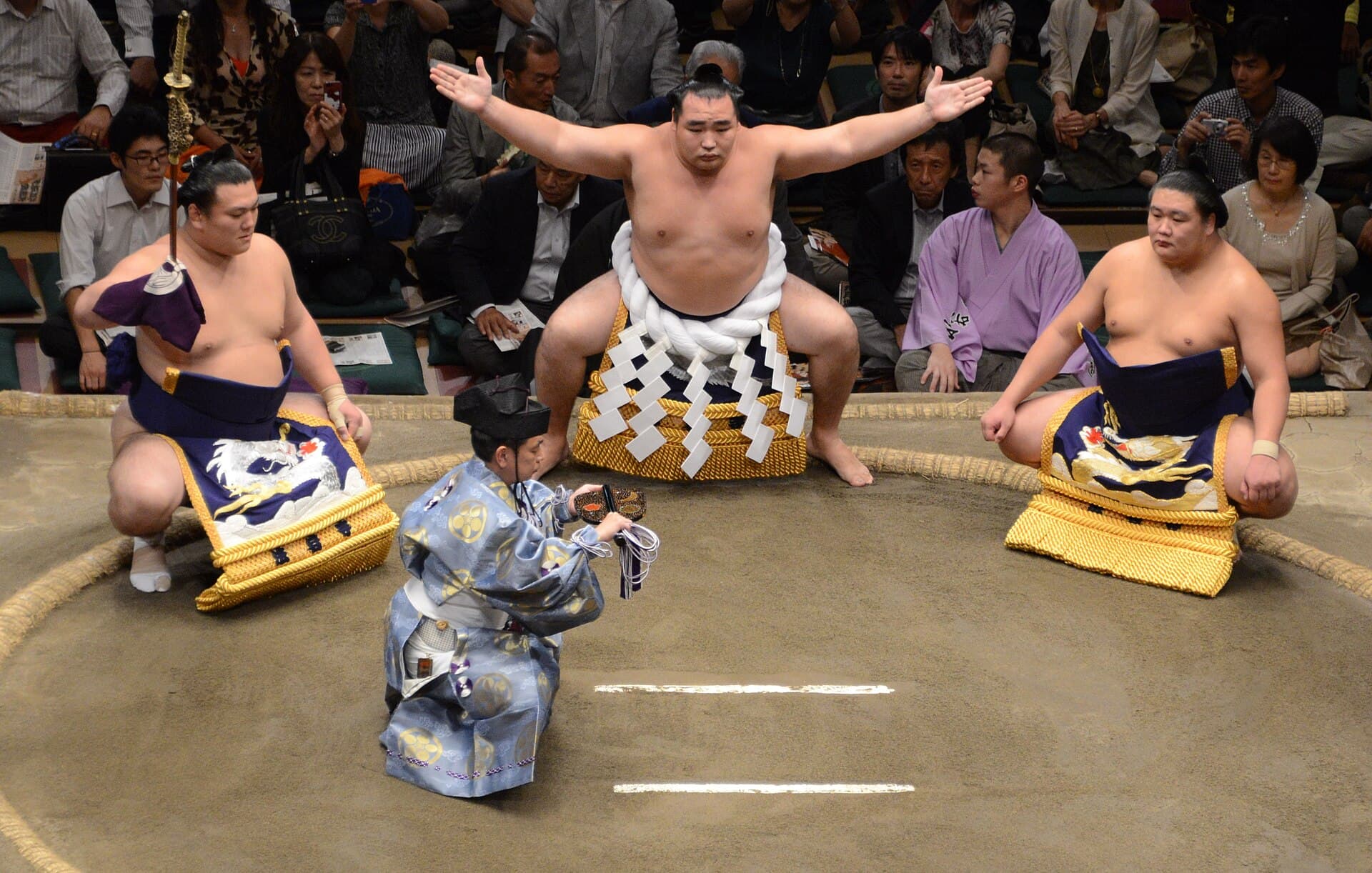 Ceremonie d'entree sur le dohyo d'un yokozuna lors du tournoi d'automne 2014 a Tokyo : le lutteur accroupi bras ecartes porte la corde de chanvre blanche, encadre par son porteur de sabre et son assistant agenouilles sur les ballots de paille