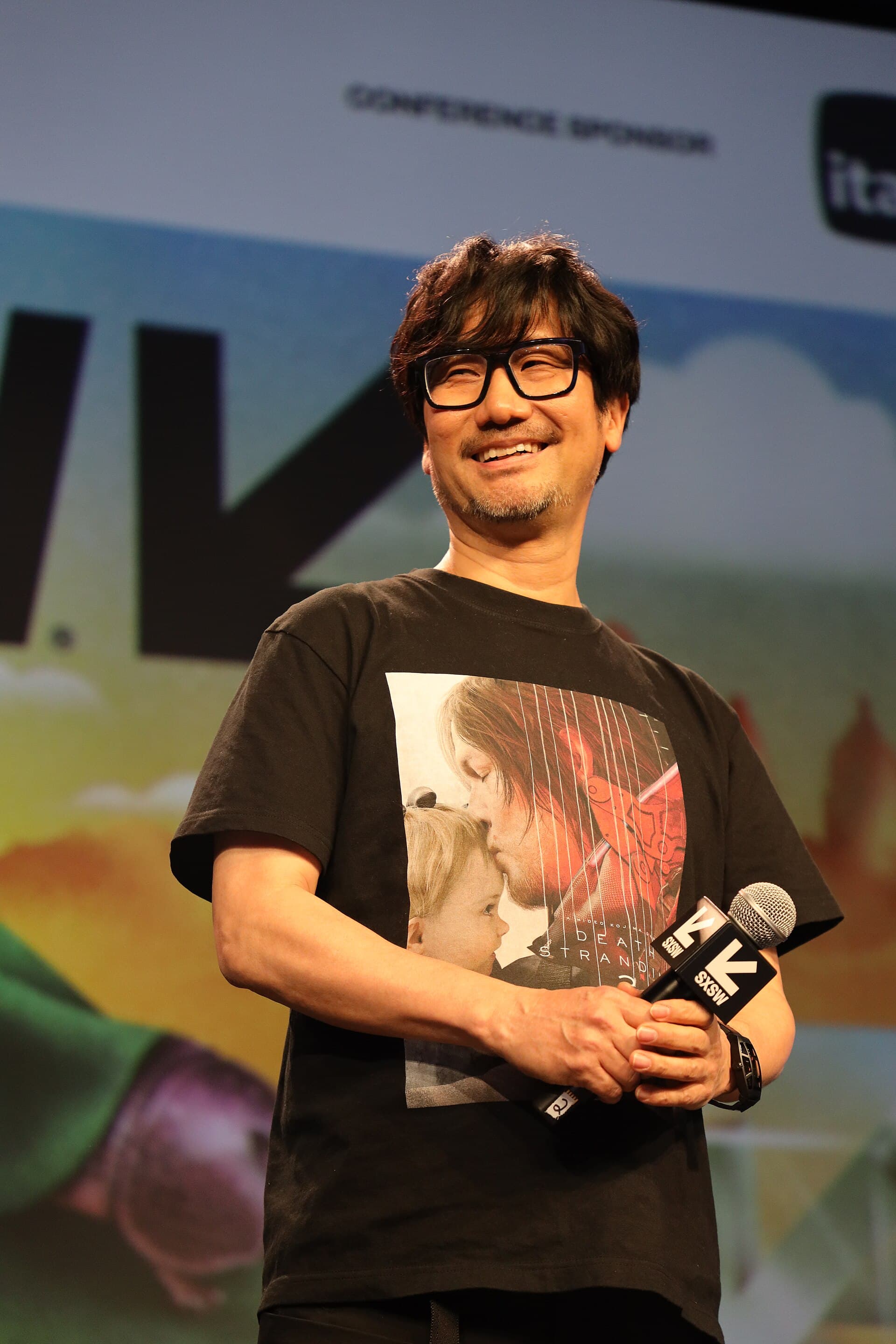 Hideo Kojima souriant sur scene au festival SXSW en 2025, micro a la main, vetu d'un t-shirt a l'effigie de Death Stranding