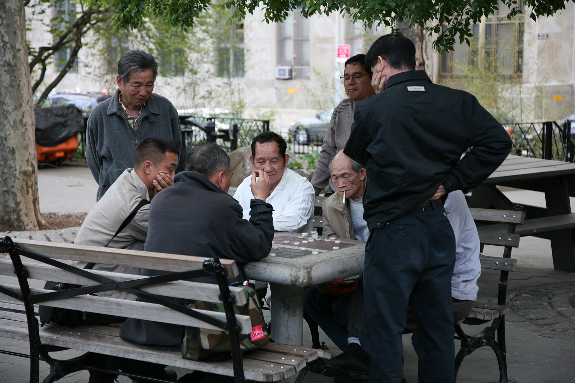 Partie de xiangqi en plein air : deux joueurs penches sur un plateau pose sur une table de parc, les pieces rondes marquees de caracteres chinois disposees sur les intersections de part et d'autre du fleuve central, des spectateurs autour de la table