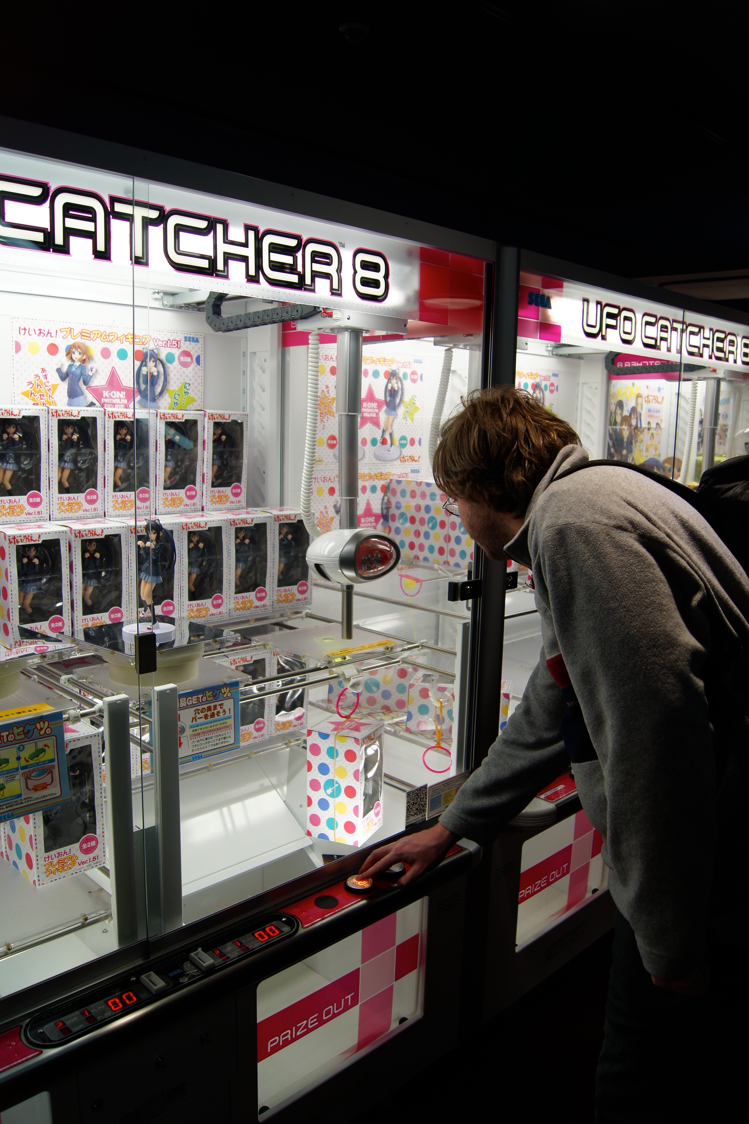 Crowded amusement arcade in Akihabara, Tōkyō, its glowing cabinets lined up in the dimness, a showcase of the Japanese gaming culture that mobile gacha inherited.